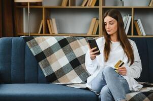 contento giovane donna Tenere Telefono e credito carta seduta su il divano. in linea acquisti. foto