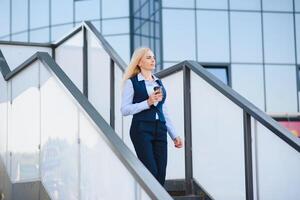 bellissimo donna andando per opera con caffè a piedi vicino ufficio costruzione. ritratto di riuscito attività commerciale donna Tenere tazza di caldo bere. foto