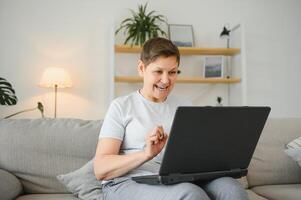 eccitato maturo donna guardare a il computer portatile schermo, lettura bene notizia nel Messaggio, festeggiare in linea lotteria vincita, gioia successo, felicissimo più vecchio femmina seduta su divano a casa, utilizzando computer. foto