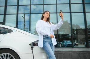 Telefono nel mani. donna su il elettrico macchine caricare stazione a giorno. marca nuovo veicolo. foto