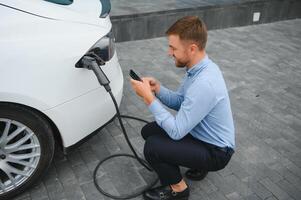 bello barbuto tipo seduta vicino il suo nuovo moderno elettrico auto e Tenere spina di il caricabatterie, mentre auto è ricarica a il ricarica stazione. foto