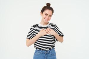 giovane sorridente donna, si sente gratitudine, grato per qualche cosa, detiene mani su cuore e sembra tenero a telecamera, in piedi al di sopra di bianca sfondo foto