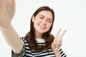 smartphone Visualizza, donna Tenere mobile Telefono telecamera e assunzione autoscatto con pace, segno v gesto, sorridente a telecamera, in posa al di sopra di bianca sfondo foto