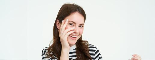 ritratto di attraente, sorridente giovane donna, ridendo e sorridente, tocchi sua viso, in piedi al di sopra di bianca sfondo foto