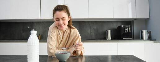 ritratto di contento giovane donna Leans su cucina piano di lavoro e mangiare cereali, ha latte e ciotola nel davanti di suo, avendo sua colazione, indossare accappatoio foto