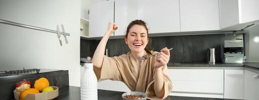 ritratto di entusiasta giovane donna mangiare cereali con latte, guardare eccitato e felice, seduta vicino cucina piano di lavoro e avendo colazione, raccolta mano su nel trionfo foto