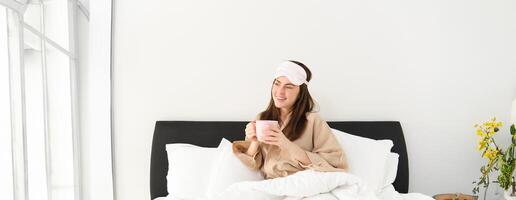 ritratto di bellissimo donna nel addormentato Borsa e pigiama, potabile caffè nel letto, godendo sua luminosa, rilassante mattina foto