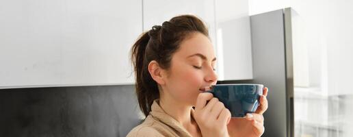 quotidiano routine e stile di vita. giovane bellissimo donna nel accappatoio, in piedi nel cucina con tazza di caffè, potabile Tè, sorridente e guardare contento foto