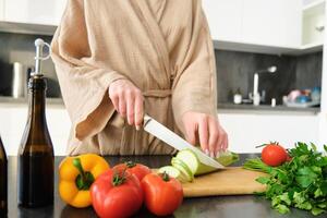 salutare stile di vita. giovane donna nel accappatoio preparazione cibo, chopping la verdura, cucinando cena su cucina contatore, in piedi al di sopra di bianca sfondo foto