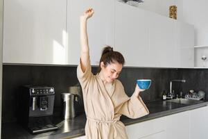 ritratto di contento ragazza danza con caffè nel il cucina, indossare accappatoio, godendo sua mattina routine foto
