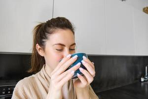 ritratto di spensierato sorridente donna potabile caffè, in piedi nel cucina con incantato, lieta viso espressione, veglia su nel il mattina e avendo cappuccino foto