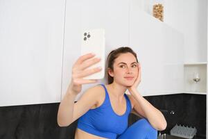 ritratto di fitness ragazza in posa per foto, assunzione autoscatto su smartphone app, seduta nel cucina, indossare abbigliamento sportivo foto