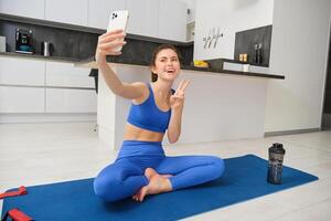 attivo giovane donna, vlogger fa gli sport, record sua allenarsi formazione a partire dal casa su smartphone telecamera, in posa per autoscatto dentro sua Casa, si siede su gomma da cancellare yoga stuoia nel blu ghette e reggiseno sportivo foto