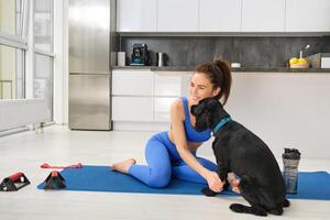 ritratto di bellissimo brunetta fitness ragazza, giochi con sua cane durante allenarsi formazione sessione, si siede a casa su gomma da cancellare yoga stuoia, fa sport esercizi nel vivente camera foto