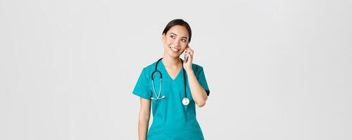 covid19, assistenza sanitaria lavoratori e prevenire virus concetto. bella sorridente asiatico femmina medico, medico nel scrub avendo conversazione, parlando su Telefono e guardare superiore sinistra angolo sognante foto