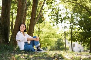 bellissimo sorridente ragazza si siede vicino albero nel parco, godendo natura all'aperto, rilassante e riposo su fresco aria foto