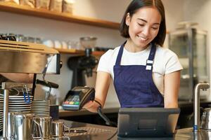 sorridente asiatico barista ragazza in lavorazione ordine, accedere ordine nel pos terminale, Lavorando a contatore nel caffè negozio foto