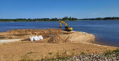 un' piccolo giallo scavatrice è Lavorando per rafforzare il Riva del fiume su un' estate giorno. foto