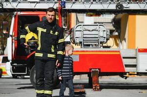 un' pompiere prendere un' poco bambino ragazzo per Salva lui. fuoco motore auto su sfondo. vigile del fuoco con ragazzo nel il suo braccia. protezione concetto. foto