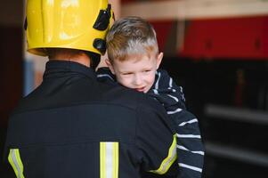 sporco pompiere nel uniforme Tenere poco salvato ragazzo in piedi su nero sfondo. foto