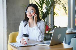 bellissimo giovane libero professionista donna utilizzando il computer portatile computer seduta a bar tavolo. contento sorridente ragazza Lavorando in linea o studiando e apprendimento mentre utilizzando taccuino. libero professionista opera, attività commerciale persone concetto. foto