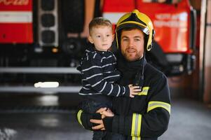 un' pompiere prendere un' poco bambino ragazzo per Salva lui. fuoco motore auto su sfondo. vigile del fuoco con ragazzo nel il suo braccia. protezione concetto. foto