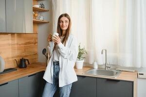 contento giovane donna Tenere un' tazza di tè mentre in piedi nel il cucina a casa nel il mattina. salutare stile di vita concetto. foto