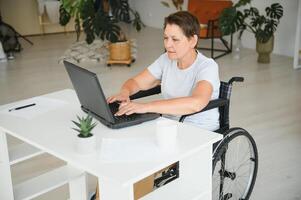 positivo anziano donna nel sedia a rotelle Lavorando su computer portatile, esso libero professionista, in linea foto