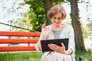 attraente maturo donna seduta su un' panchina Tenere e utilizzando un' digitale tavoletta nel il parco a estate giorno foto