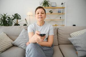 di mezza età donna seduta su il divano e svolta su il tv con un' a distanza controllo. foto