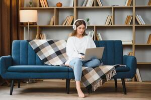 sorridente attraente giovane donna seduta su divano utilizzando il computer portatile comunicare Lavorando in linea a casa, contento adolescente ragazza digitando su computer, godendo scrittura blog o chat con amici nel sociale Rete foto