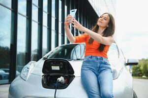 giovane donna è in piedi vicino il elettrico auto e sembra a il inteligente Telefono. il noleggio auto è ricarica a il ricarica stazione per elettrico veicoli. auto condivisione. foto