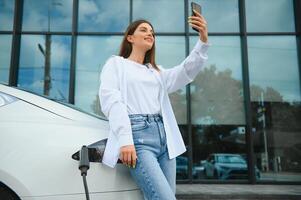 donna con Telefono vicino un noleggio elettrico macchina. veicolo carico a il ricarica stazione foto