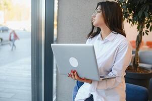 bellissimo caucasico donna sognare di qualcosa mentre seduta con portatile netbook nel moderno bar sbarra, giovane affascinante femmina libero professionista pensiero di nuovo idee durante opera su il computer portatile computer foto