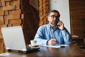 positivo anziano barbuto uomo con grigio capelli potabile caffè e utilizzando il computer portatile a bar, copia spazio. elegante anziano uomo d'affari nel Borgogna giacca godendo il suo tè mentre Lavorando in linea. foto