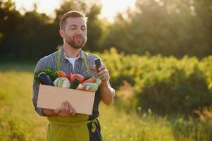 contadino Tenere un' gabbia di bio verdure nel il azienda agricola. contento uomo mostrando scatola di raccolto verdure. foto