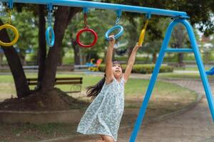 contento ragazza sospeso su scimmia bar di mano fare esercizio. poco asiatico ragazza giocando a all'aperto terreno di gioco nel il parco su estate vacanza. salutare attività. foto