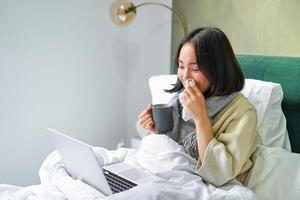 laborioso asiatico ragazza soggiorno nel letto con freddo, attraente influenza ma Lavorando a partire dal casa con computer portatile, studiando mentre sensazione malato foto
