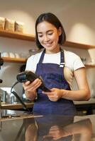 bellissimo sorridente asiatico ragazza, barista inserire cliente credito carta nel pos terminale, in lavorazione pagamento, assunzione ordini nel caffè negozio foto