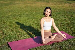 consapevolezza e meditazione. giovane asiatico donna sorridente mentre fare yoga, rilassante nel asana su gomma da cancellare stuoia, fare esercizi nel parco su fresco aria foto