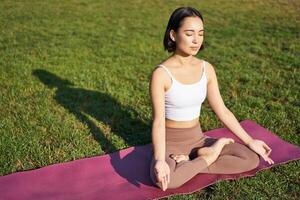 consapevolezza e meditazione. giovane asiatico donna sorridente mentre fare yoga, rilassante nel asana su gomma da cancellare stuoia, fare esercizi nel parco su fresco aria foto