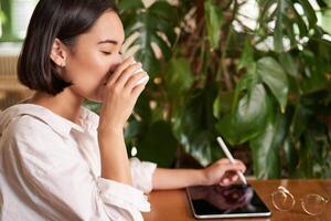 bellissimo asiatico ragazza seduta nel bar con tazza di caffè, disegno su digitale tavoletta con grafico penna, scherzetto e fabbricazione graffi foto