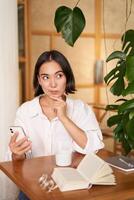 bellissimo giovane donna, 25 anni vecchio, pensiero, Tenere smartphone e guardare premuroso, seduta nel bar, decidere smth foto