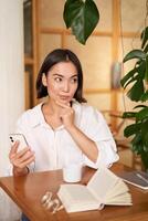 bellissimo giovane donna, 25 anni vecchio, pensiero, Tenere smartphone e guardare premuroso, seduta nel bar, decidere smth foto