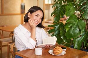 sognante giovane sorridente asiatico donna lettura prenotare, seduta nel bar, mangiare brioche e potabile caffè nel accogliente interno foto
