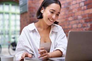 giovane sorridente asiatico donna con senza fili auricolari, seduta nel coworking ufficio spazio con computer portatile, fare compiti a casa, fabbricazione Appunti foto