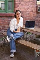 verticale tiro di bella vista asiatico donna con computer portatile, seduta nel all'aperto bar, potabile caffè e sorridente, ridendo spensierato foto