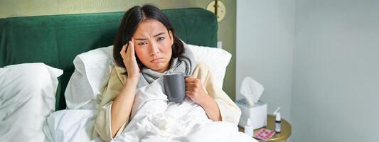 ritratto di asiatico donna con male alla testa, attraente col, soggiorno su malato partire a casa, dire bugie nel letto, potabile caldo Tè, avendo influenza foto