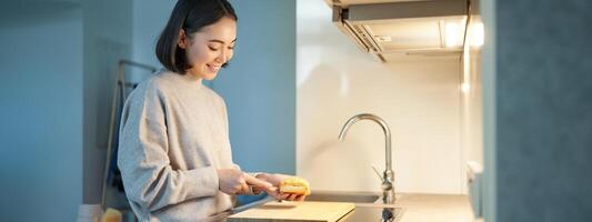 ritratto di sorridente giovane asiatico donna in piedi su cucina e fabbricazione un' panino, cucinando per se stessa foto