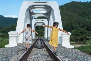 bambini giocando al di fuori su treno brani. asiatico sorelle a piedi su il ferrovia nel rurale scena. foto